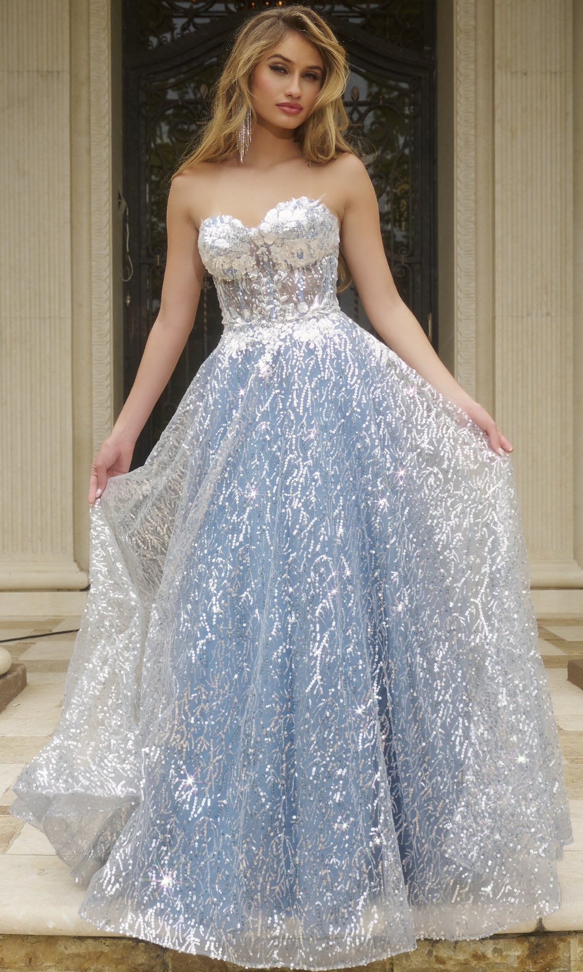 A woman stands before a building, gracefully holding out the sides of her Strapless Long Sequin Prom Ball Gown: Jovani 08417—a blue and silver sparkling sweetheart dress.